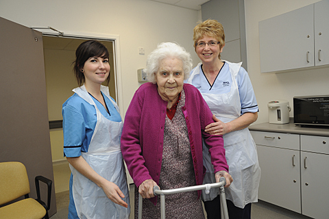 Two carers either side of an older lady assisting her to walk with her walking frame