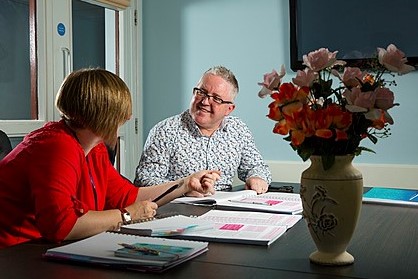 Man and woman sitting at table with papers spread in front of them talking, there is also a vase of flowers on the table and a tv screen behind them