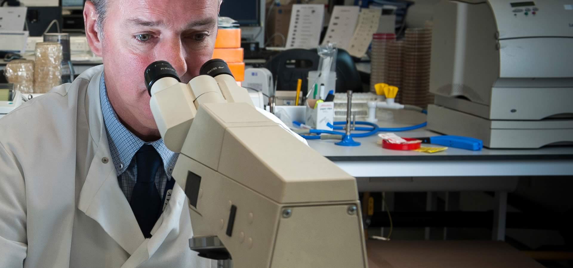 A man in a white coat sitting at a microscope placing a slide under the viewer