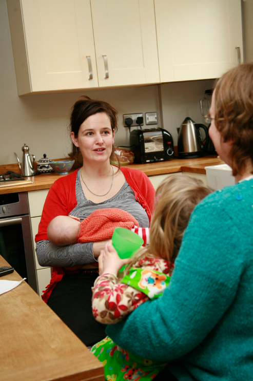 New mum breastfeeding baby in kitchen while talking to another mum