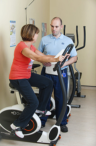 Patient using gym bike while physio talks to her.