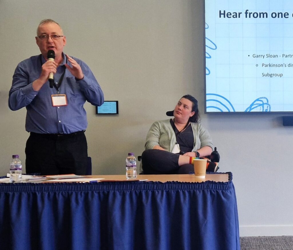 A man with a blue shirt standing up and talking into a microphone giving a presentation