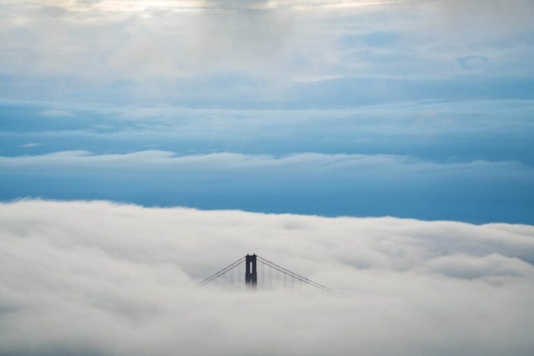 The tallest part of a suspension bridge peeking through thick white clouds, as seen from a high vantage point
