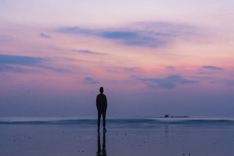 A silhouette of a man on a wet beach looking out to a pink evening sky